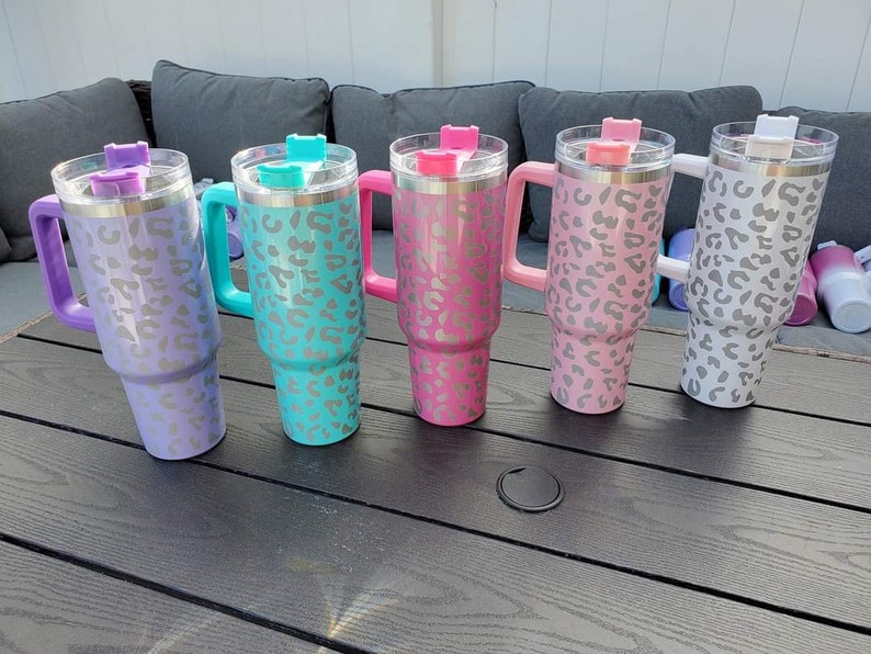 May include: Five stainless steel travel mugs with a leopard print design. The mugs are in different colors: purple, teal, pink, light pink, and white. Each mug has a handle and a lid.