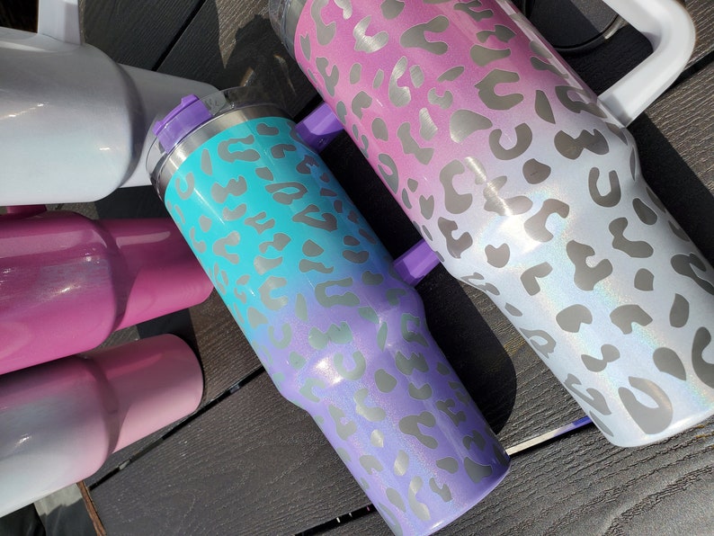 May include: Three stainless steel tumblers with a leopard print design. The tumblers are ombre, with a light blue, pink, and white color gradient.