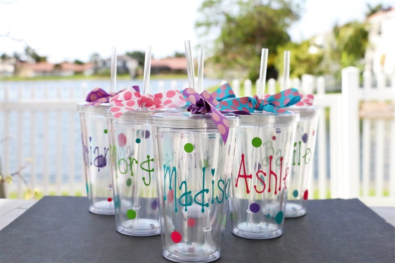 Op de afbeelding: Zes doorzichtige plastic bekers met rietjes en kleurrijke stippenboogjes. Op elke beker staat een naam: "Brian", "Lori", "Madison", "Ashlie", "Bailie" en "Olivia".