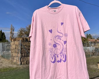 May include: A light pink t-shirt with a purple graphic of a snake, hearts, and flowers. The word "uwu" is printed in purple below the snake. The t-shirt is hanging on a pink hanger against a blue sky.