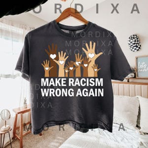May include: Dark grey t-shirt featuring a graphic of hands in diverse skin tones, each holding a white heart. The text "MAKE RACISM WRONG AGAIN" is printed in white below the hands. The t-shirt is suspended from a wooden hanger.