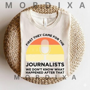 May include: Cream-colored t-shirt with a graphic featuring a microphone and the text "FIRST THEY CAME FOR THE JOURNALISTS WE DON'T KNOW WHAT HAPPENED AFTER THAT." The design incorporates orange, yellow, and white colors, set against a woven background.