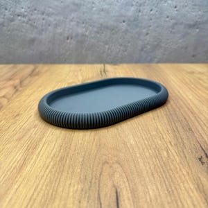 Könnte beinhalten: Ein oval geformtes Tablett in einem einfarbigen Schieferblau. Das Tablett verfügt über eine glatte, flache Innenfläche und einen erhöhten, dicken Außenrand, der mit einer durchgehenden vertikalen Rippenstruktur versehen ist.