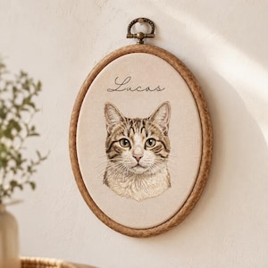 May include: An oval wooden embroidery hoop frame containing a beige fabric panel with a detailed stitched portrait of a brown tabby cat. The name Lucas is embroidered in black cursive script above the cat. A metal hanging loop is at the top.