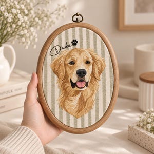May include: An oval wooden embroidery hoop containing fabric with a vertical light green and white striped pattern. The fabric features an embroidered golden retriever portrait, the name Duke in black script, and a small black paw print.