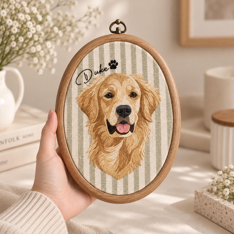 May include: An oval wooden embroidery hoop containing fabric with a vertical light green and white striped pattern. The fabric features an embroidered golden retriever portrait, the name Duke in black script, and a small black paw print.