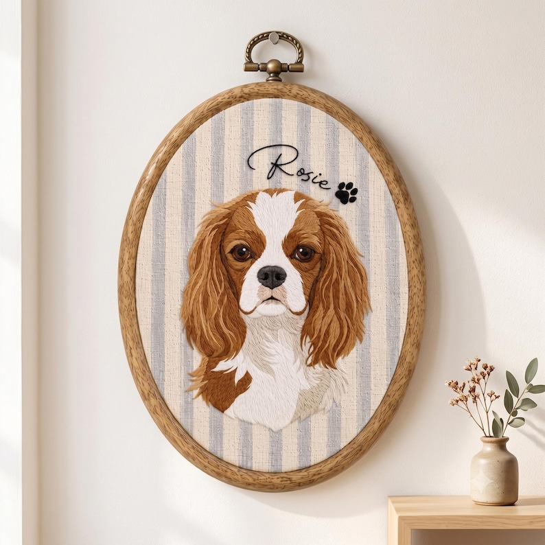May include: An oval wooden embroidery hoop frame containing a fabric portrait of a brown and white Cavalier King Charles Spaniel. The fabric has a light blue and white vertical stripe pattern. The name Rosie and a paw print are stitched above.
