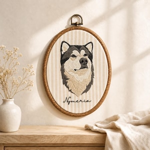 May include: An oval embroidery hoop featuring a detailed stitched portrait of a husky dog with black, gray, and white fur. The fabric background has vertical beige stripes, and the name Nymeria is embroidered in black script below the dog.