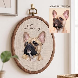 May include: An oval wooden embroidery hoop containing a custom fabric portrait of a tan French Bulldog with a black muzzle. The name Lucy is embroidered in black script above the dog. A small inset shows a reference photo of the same dog.
