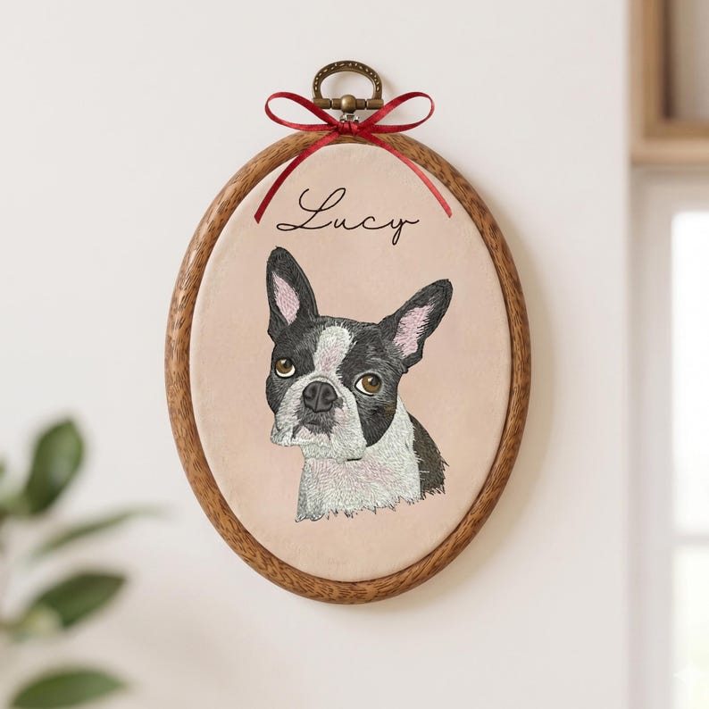May include: An oval wooden embroidery hoop containing a fabric panel with a stitched portrait of a black and white Boston Terrier dog. The name Lucy is embroidered in black script above the dog. A red ribbon is tied at the top metal hardware.