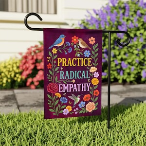 May include: A garden flag with the words "PRACTICE RADICAL EMPATHY" in colorful text. The flag features illustrations of birds, flowers, and a hummingbird against a deep purple background. The flag is displayed on a black metal stand in a garden setting.