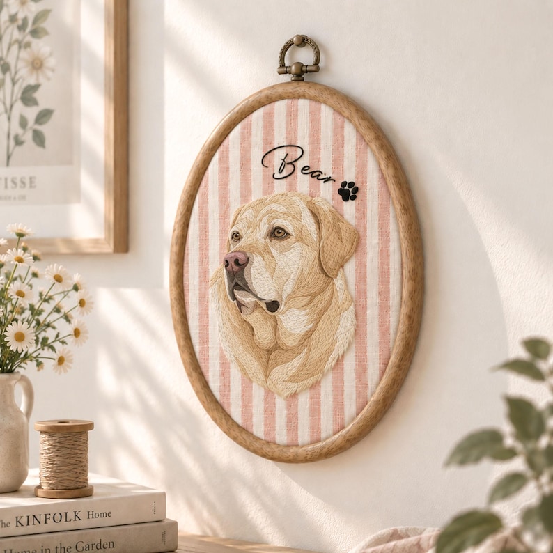 May include: An oval wooden embroidery hoop containing fabric with vertical pink and white stripes. The fabric features an embroidered portrait of a yellow Labrador dog with the text Bear and a small black paw print icon above the dog.
