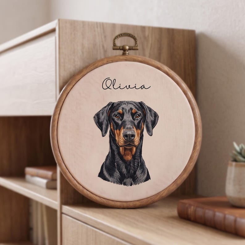 May include: A circular wooden embroidery hoop frame containing a light beige fabric panel. The fabric features a detailed illustration of a black and tan Doberman dog head centered below the black cursive text that reads Olivia.