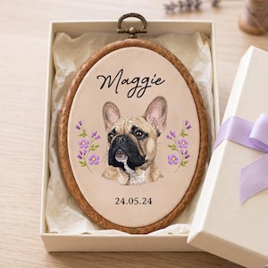 May include: An oval wooden embroidery hoop frame containing a fabric insert with a printed illustration of a fawn French Bulldog. The name Maggie is printed above the dog, and the date 24.05.24 is printed below, flanked by purple floral sprigs.