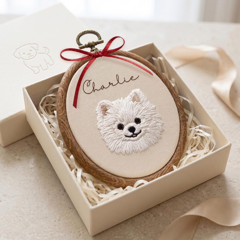 May include: An oval embroidery hoop featuring a white fabric panel with a stitched portrait of a white dog and the name Charlie in black script. The hoop has a dark wood grain frame, a bronze metal hanging loop, and a red ribbon tied at the top.