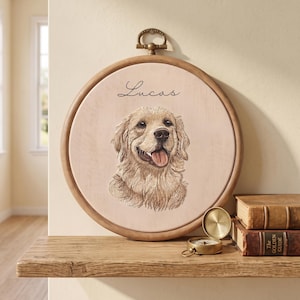May include: A circular wooden embroidery hoop frame containing a light-colored fabric panel with a detailed embroidered portrait of a golden retriever dog and the name Lucas stitched in black script above the dog's head.