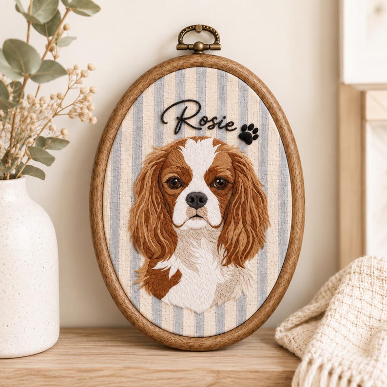 May include: An oval wooden embroidery hoop containing a fabric portrait of a Cavalier King Charles Spaniel. The dog has brown and white fur. Above the dog, the name Rosie is embroidered in black script, followed by a small black paw print.