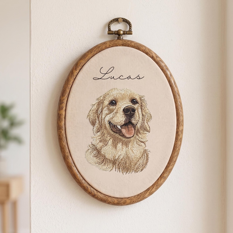 May include: An oval wooden embroidery hoop frame containing a beige fabric panel with a detailed thread-stitched portrait of a golden retriever dog. The name Lucas is embroidered in black script above the dog. A metal hanging loop is at the top.