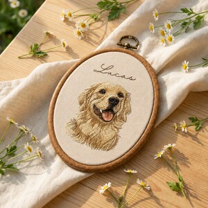 May include: An oval wooden embroidery hoop containing a cream-colored fabric panel with a detailed thread-stitched portrait of a golden retriever dog and the name Lucas embroidered in black cursive script above the dog's head.