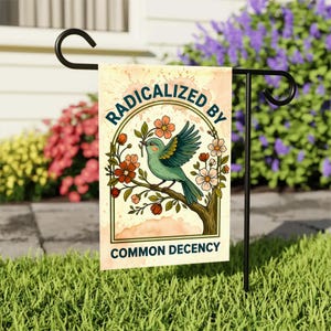 May include: A garden flag with a teal bird perched on a tree branch, surrounded by flowers. The flag reads "RADICALIZED BY COMMON DECENCY" in a circular design. The flag is displayed on a black metal stand in a garden setting.