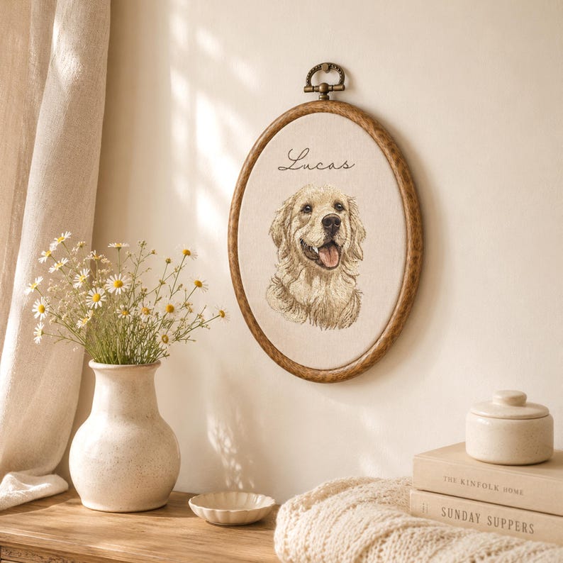 May include: An oval wooden embroidery hoop containing a cream-colored fabric panel with a detailed stitched portrait of a golden retriever dog and the name Lucas embroidered in black script above the dog's head, featuring a metal hanging loop.