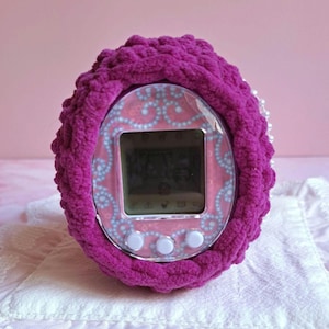 Puede incluir: Una funda de ganchillo magenta rodea un juego digital de mascotas portátil vintage. El juego tiene una cara rosa con un patrón azul y blanco, una pequeña pantalla y tres botones blancos. La funda está hecha de hilo grueso y texturizado.
