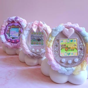 Peut inclure: Trois jeux électroniques pour animaux de compagnie aux couleurs pastel, ornés de bordures et de nœuds en peluche. Ils reposent sur des socles blancs en forme de nuage sur un fond rose. Les écrans affichent des scènes pixelisées d'animaux virtuels.