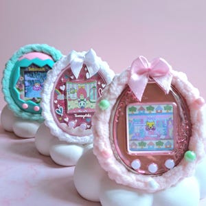 Peut inclure: Trois Tamagotchi aux couleurs pastel, chacun avec une base en forme de nuage et un cadre en crochet duveteux. Les écrans affichent des personnages animés dans un monde virtuel. Les appareils sont roses, vert menthe et blancs.