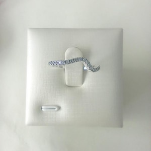 May include: A silver-colored ring with a wave design, set with small, clear gemstones. The ring is displayed on a white, textured surface, showcasing its curved shape and sparkling details. The ring's band is thin and delicate.