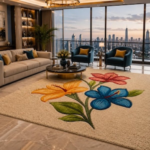 Op de afbeelding: Beige vloerkleed met een groot bloemmotief in oranje, rood en blauw. Het vloerkleed ligt in een moderne woonkamer met een bank, fauteuils en uitzicht op de stad door grote ramen.
