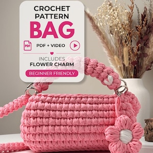 May include: A pink crochet bag with a thick braided handle and a matching flower charm. The text on the overlay reads: CROCHET PATTERN BAG, PDF + VIDEO, INCLUDES FLOWER CHARM, BEGINNER FRIENDLY. The bag features a textured stitch pattern.