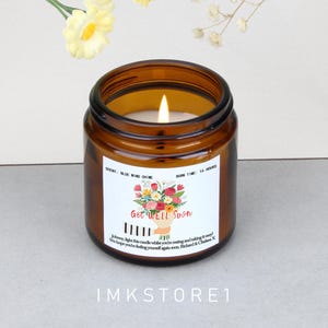 Può includere: Un barattolo di candela in vetro marrone acceso con un'etichetta bianca che recita "Get Well Soon" con un disegno floreale. La candela ha un tempo di combustione di 16 ore e una fragranza "Blue Wind Chime". Il testo "IMKSTORE1" è in basso.