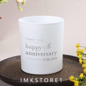 May include: A white cylindrical candle holder with the text "Connor + Leia happy 5th anniversary 17.06.2019" in gray. The candle holder sits on a dark brown plate, with yellow flowers in the background. The shop's name, IMKSTORE1, is at the bottom.