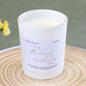 May include: A white candle in a glass jar with a label that reads "to my Bridesmaid on my Wedding day." The candle has a burn time of 15 hours. The candle is on a wooden surface.