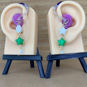 May include: A pair of earrings with a gold chain, white and green star charms, and a small white bead. The earrings are displayed on a beige ear model with a purple and clear earbud.
