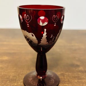 May include: A ruby red wine glass with a decorative design. The glass features a detailed etched pattern of swirls and a landscape scene. The stem and base are also red, creating a cohesive and elegant look. The glass is likely made of crystal.