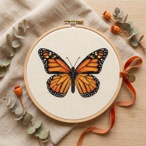 May include: A completed cross-stitch project of a monarch butterfly, displayed in a wooden embroidery hoop. The butterfly features orange and black wings with white-dotted edges. A rust-colored ribbon and dried floral elements surround the hoop.