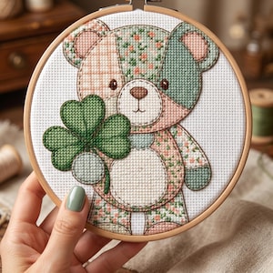 Puede incluir: Un bastidor de bordado circular de madera que contiene un patrón de punto de cruz de un oso de peluche. El oso está compuesto por secciones de patchwork en verde salvia, rosa suave y motivos florales, sosteniendo un trébol de cuatro hojas.