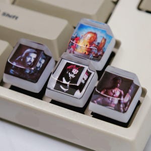 May include: A computer keyboard with custom keycaps. The keycaps display various images, including portraits and graphic designs. The keycaps are transparent with the images visible beneath the surface, and are set on a white keyboard.