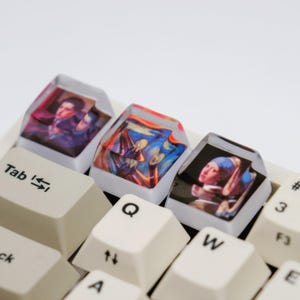 May include: Custom keyboard keycaps featuring famous art prints. The keycaps display images of classic paintings, including a portrait, "The Scream," and "Girl with a Pearl Earring." The keycaps are clear with the art visible beneath.
