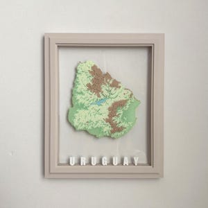 May include: A framed three-dimensional topographic map of Uruguay featuring layered green and brown terrain with a blue water feature. The word URUGUAY is displayed in white block letters at the bottom inside the light-colored frame.