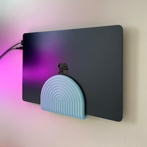 Pode incluir: Um portátil preto mate está montado verticalmente contra uma parede usando um suporte de parede azul claro em forma de semicírculo. O suporte apresenta uma série de cristas concêntricas em relevo em forma de arco, criando uma superfície estriada.