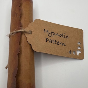 Pode incluir: Um rolo de papel castanho enrolado, amarrado com barbante, acompanhado por uma etiqueta castanha. A etiqueta diz "Hypnotic Pattern" em escrita preta e tem recortes em forma de coração. O pergaminho tem uma aparência envelhecida.