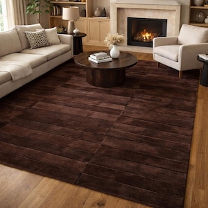 May include: A rectangular, dark brown area rug with a textured, linear design. The rug is placed on a hardwood floor in a living room setting, with a sofa, armchair, and coffee table visible in the background.