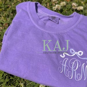Puede incluir: Una camiseta lavanda con iniciales bordadas y un diseño de lazo. La camiseta presenta el texto "KAJ SOUTHERN DESIGNS" en verde y blanco. La camiseta está doblada y sobre hierba verde.