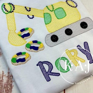 Op de afbeelding: Wit T-shirt met een geborduurde gele graafmachine, groene accenten en drie kleurrijke ringen. De naam "RORY" is geborduurd in blauw, groen en geel. "KAJ SOUTHERN DESIGNS" is in het wit gedrukt.