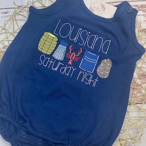 Puede incluir: Romper de bebé azul marino con texto y gráficos bordados. El texto dice "Louisiana Saturday Night" con imágenes de cangrejos de río, maíz y otros alimentos. El romper tiene cierres a presión y detalles de botones en los hombros.