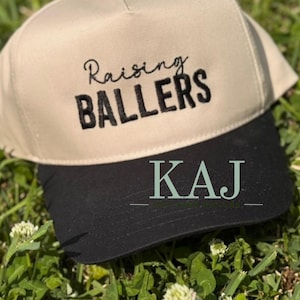 Op de afbeelding: Een beige en zwarte baseballcap met de tekst "Raising Ballers" in zwart schrift. De onderkant van de cap heeft de initialen "KAJ" in een lichtgroene kleur. De cap ligt op groen gras.