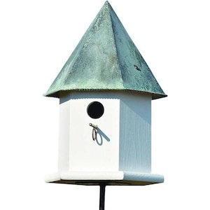 May include: A white hexagonal birdhouse featuring a pointed, weathered green copper roof. The structure includes a circular entrance hole and a small metal perch extending from the front panel, mounted on a thin black pole against a white background.