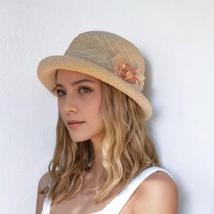 May include: A beige cloche hat with a woven texture and a small, curved brim. The hat is adorned with a cluster of fabric flowers in shades of peach and tan. The hat is a stylish accessory.